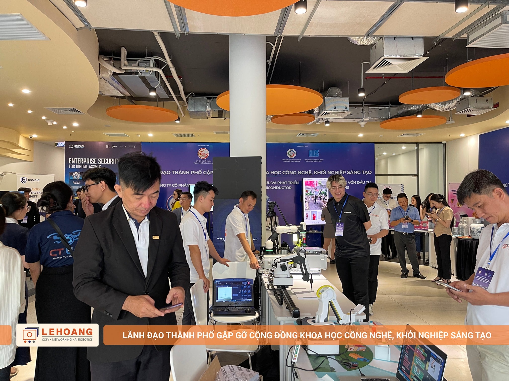 Hai nhân viên Lê Hoàng Robotics trao đổi và giới thiệu robot cánh tay Dobot cho khách tham quan tại gian hàng, bàn trưng bày các thiết bị robot giáo dục và công nghiệp, backdrop Lê Hoàng Robotics phía sau, không gian triển lãm với đèn trang trí cam