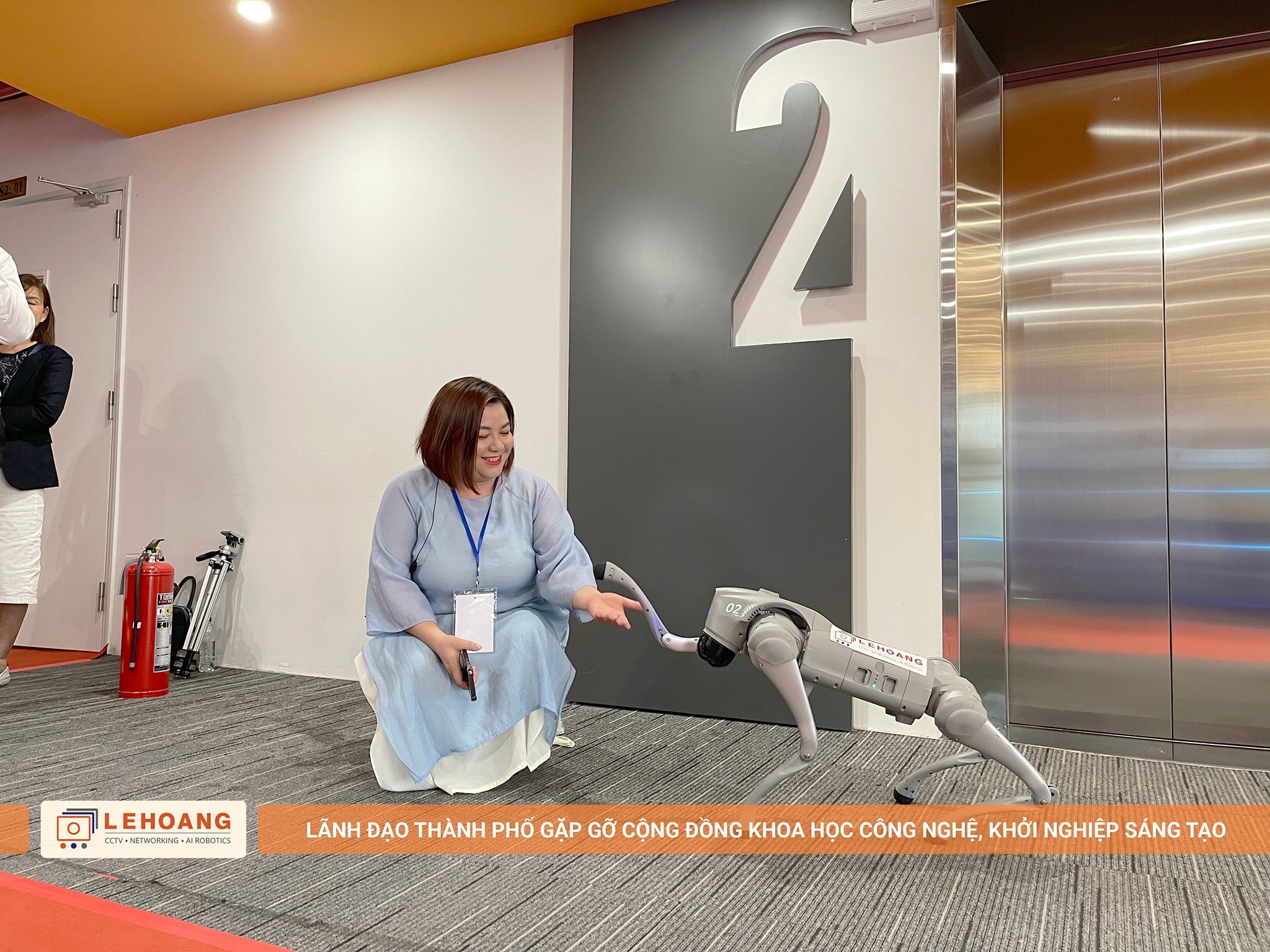 Khách tham dự ngồi xuống tương tác thân thiện với robot chó Unitree Go2, robot đang thực hiện động tác bắt tay, không gian hành lang sự kiện với số tầng 