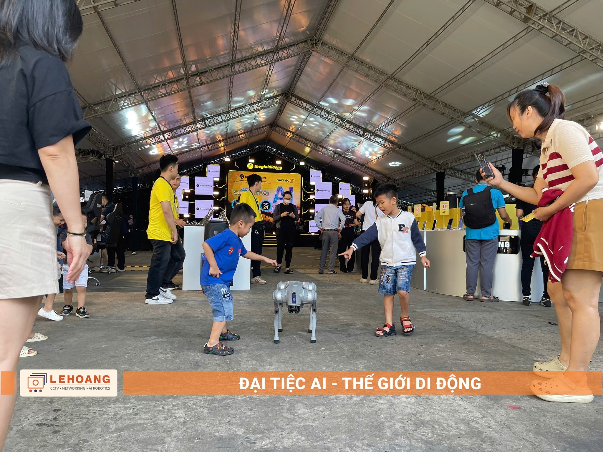  Hai bé trai hào hứng chạy theo robot Unitree Go2 trong hội trường Đại tiệc AI, phụ huynh đứng xung quanh quay video, sân khấu chính với màn hình LED ở phía xa